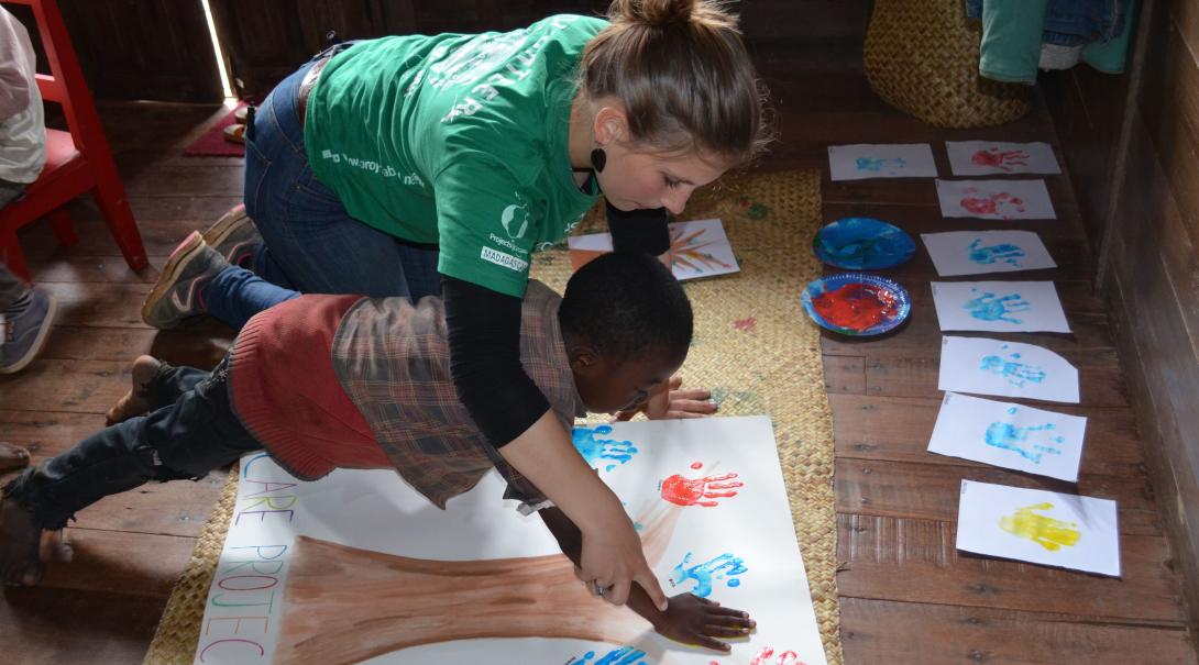 A volunteer on a mission trip to Madagascar, Africa helps a child with painting a tree. On a mission trip to Africa for high school students, a volunteer helps a child on an art project.